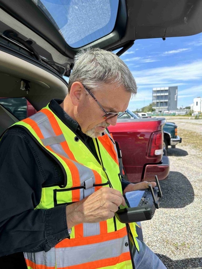 Older man in a high-visibility vest using a remote controller outdoors near a red pickup truck, blue sky above.